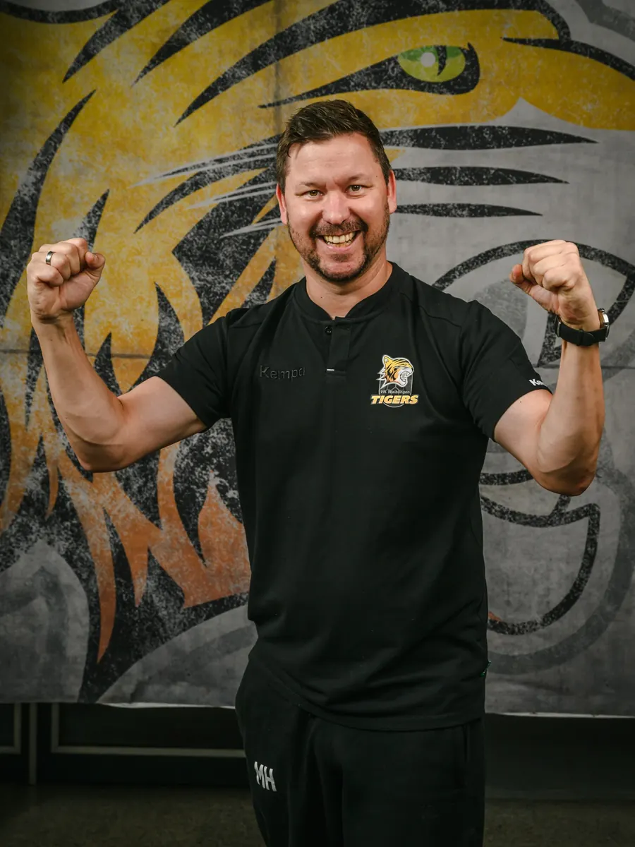 Maik im schwarzen Polo der Waiblingen Tigers reckt beide Fäuste und lacht, hinter ihm ein großflächig gemaltes Tigermotiv.
